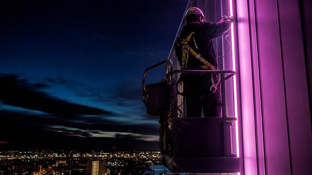 Lighting technician on an articulating boom lift connecting data cable to LED facade lighting on a tall building at dusk